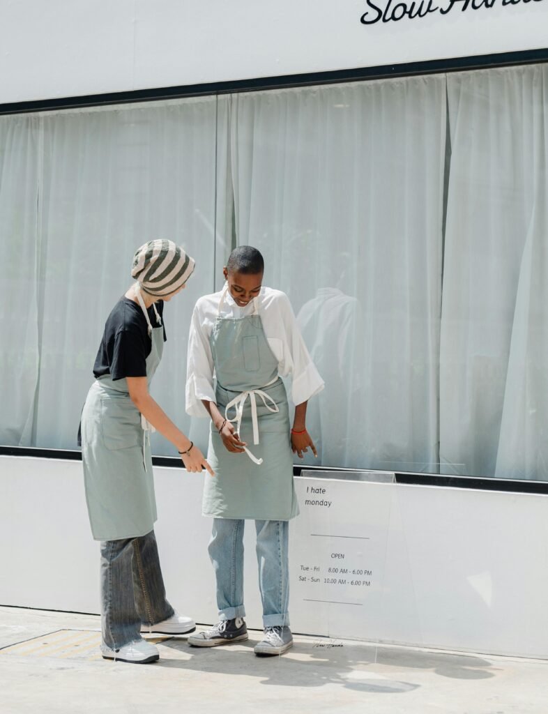 Two colleagues wearing aprons conversing in front of a cafe window during the day.